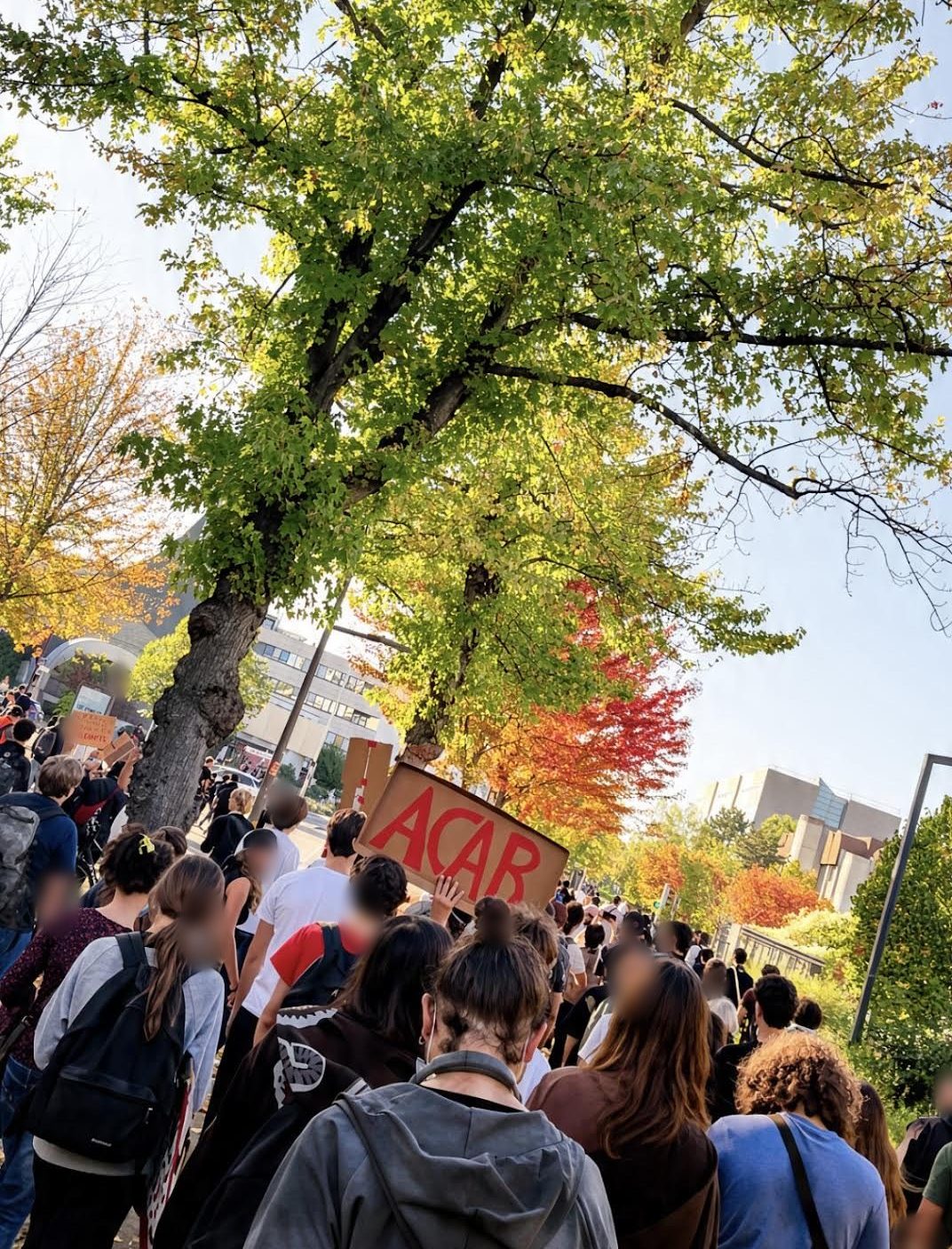 Le cortège vu de dos sous de grands arbres aux feuilles d'automne, une pancarte en carton « ACAB » levée au-dessus des têtes, des bâtiments modernes à l'arrière-plan.