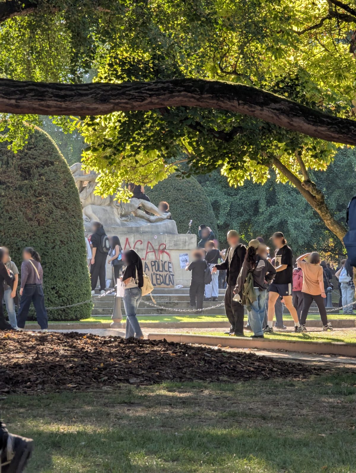 Le monument aux morts de Strasbourg vu de loin, sous les branches d'un arbre : des jeunes assis sur la sculpture, le socle tagué à la peinture rouge, une banderole en carton, des manifestants sur les marches.
