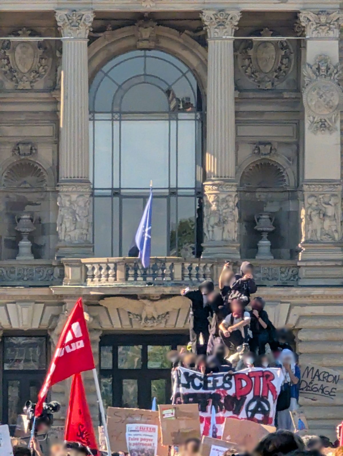 Sur le porche du Palais du Rhin, des jeunes vêtus de noir, capuches et visages masqués, derrière une banderole lycéenne marquée d'un A cerclé ; à droite, un tag « Macron dégage » ; drapeaux rouges et pancartes au premier plan.