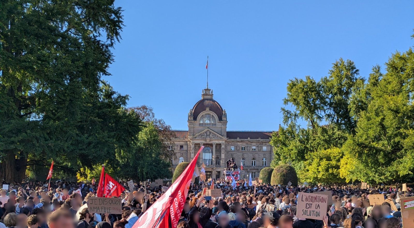 La place de la République à Strasbourg remplie de manifestants devant le Palais du Rhin, le 6 octobre 2026 : drapeaux rouges et pancartes, dont « Je dis quoi à ma fille » et « L'insoumission est la solution ».