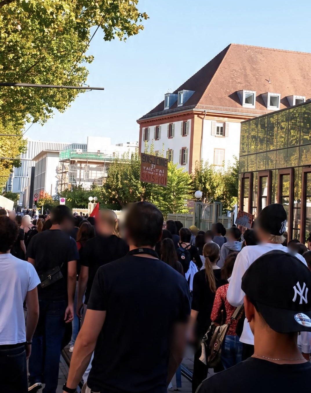 Le cortège vu de dos dans une rue bordée d'arbres ; au-dessus des têtes, une pancarte en carton où « Sois jeune et tais-toi » est corrigé : « tais-toi » barré, « bats-toi » écrit en rouge.