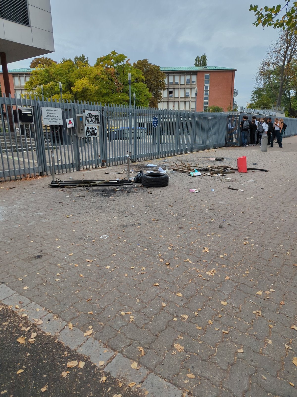 Le parvis du lycée Kléber à Strasbourg après le blocage du 1er octobre 2026 : traces de feu, pneu, branchages, grille taguée et élèves au portail.
